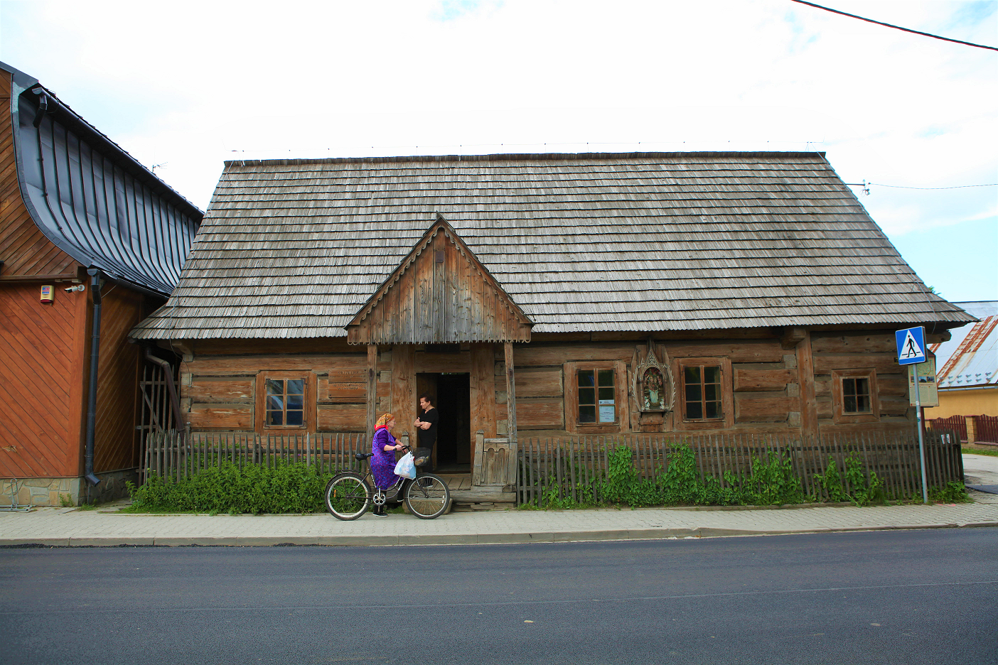 Drewniany dom od frontu, dach pokryty gontem. Przed domem dwie osoby jedna stoi, druga jedzie na rowerze po chodniku. 